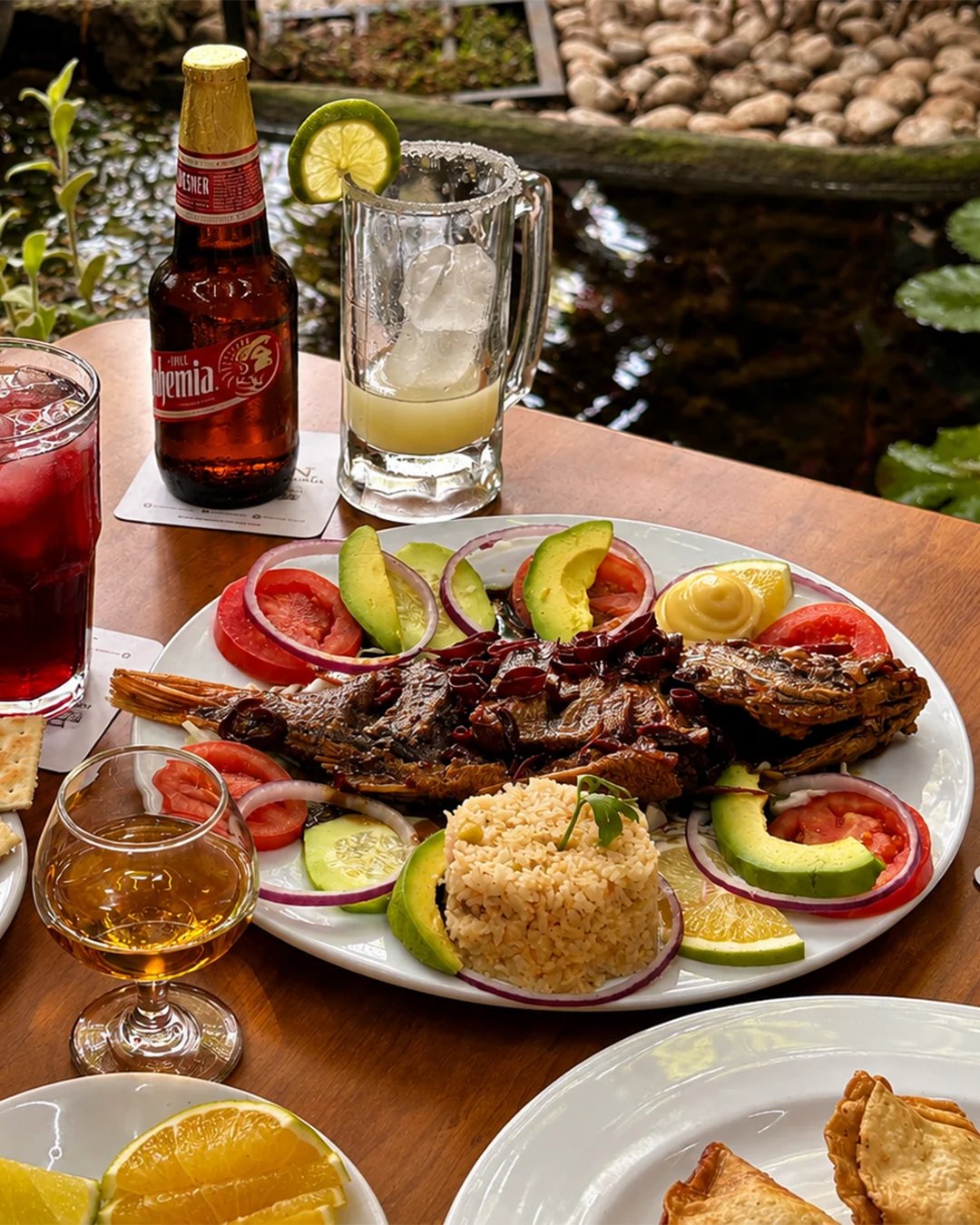 Mesa al aire libre con pescado a la talla, aguacates, cervezas y cocteles junto al estanque del jardín